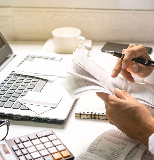 Close-up of hands flipping through financial documents and receipts on a desk, surrounded by a laptop, calculator, and notebook, representing Business One Tax & Accounting’s meticulous approach to financial management.